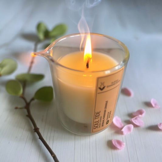 Massage Candle in a glass jar with a label, surrounded by pink petals and green leaves on a light surface.
